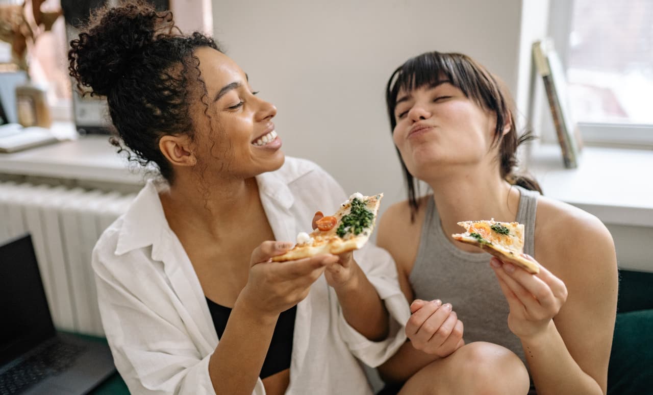 Due amiche ridono mangiando insieme una pizza, senza pensare a mangiare come una persona normale