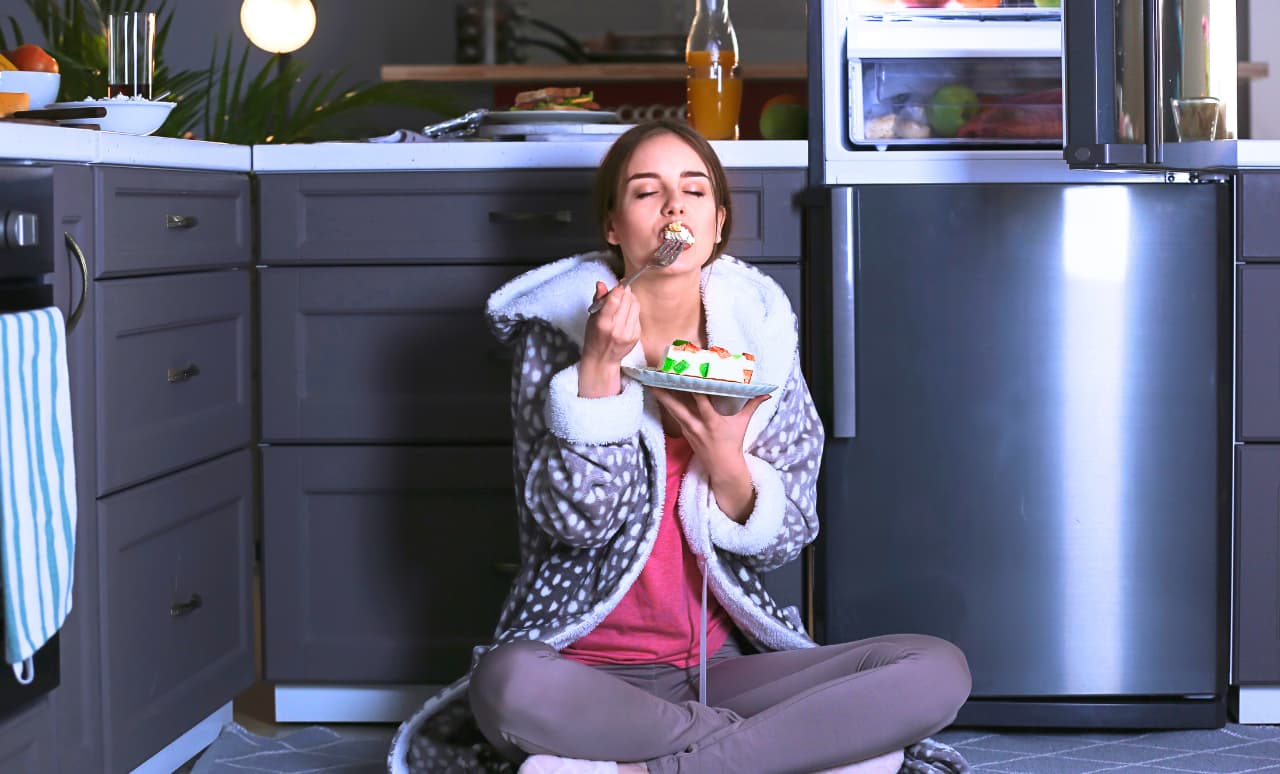 Donna seduta a terra in cucina la sera con un piatto in mano, davanti al frigorifero aperto, mentre mangia in un momento di fame emotiva